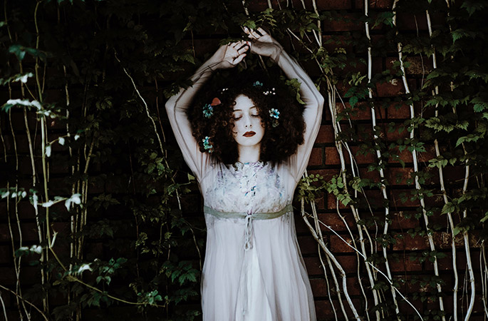 Woman with flowers in her hair and hands in the air standing against a brick wall covered in greenery.