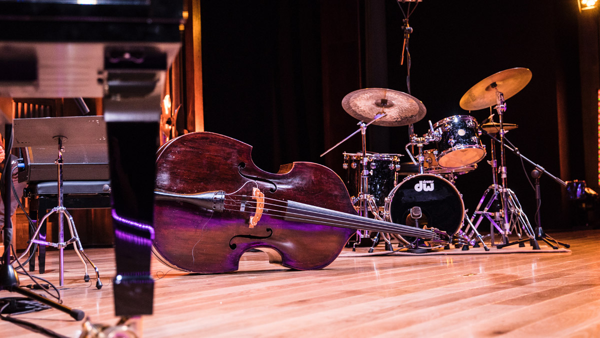 A polished wooden stage with a paino in the foreground, cello in the midground, and drum kit in the background. A black curtain sits behind the stage.