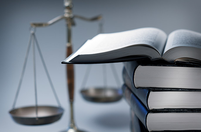 Scales of justice in the background and a stack of books in the foreground. The top book is open.