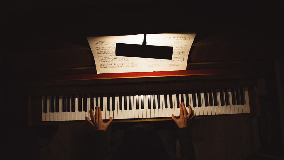 Birds-eye view of a person playing an upright piano in a dark room. A music book is open in front of them.