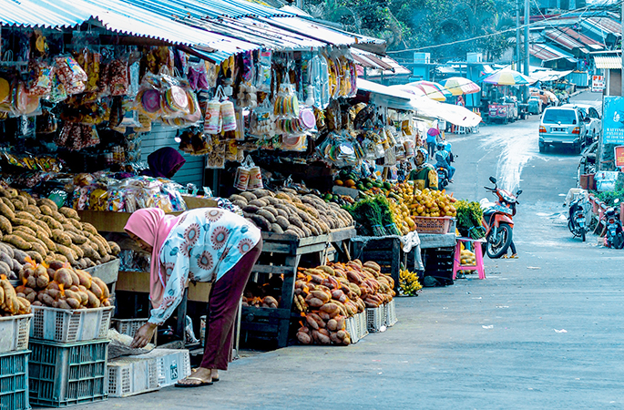 An Indonesian food market next to a road. A woman is browsing through a shop in the foreground.