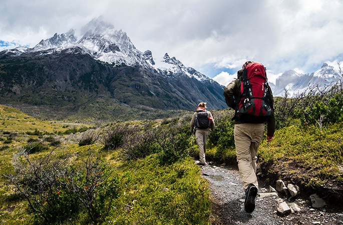 Two people hiking along a dirt path with snow-capped mountains in the background.